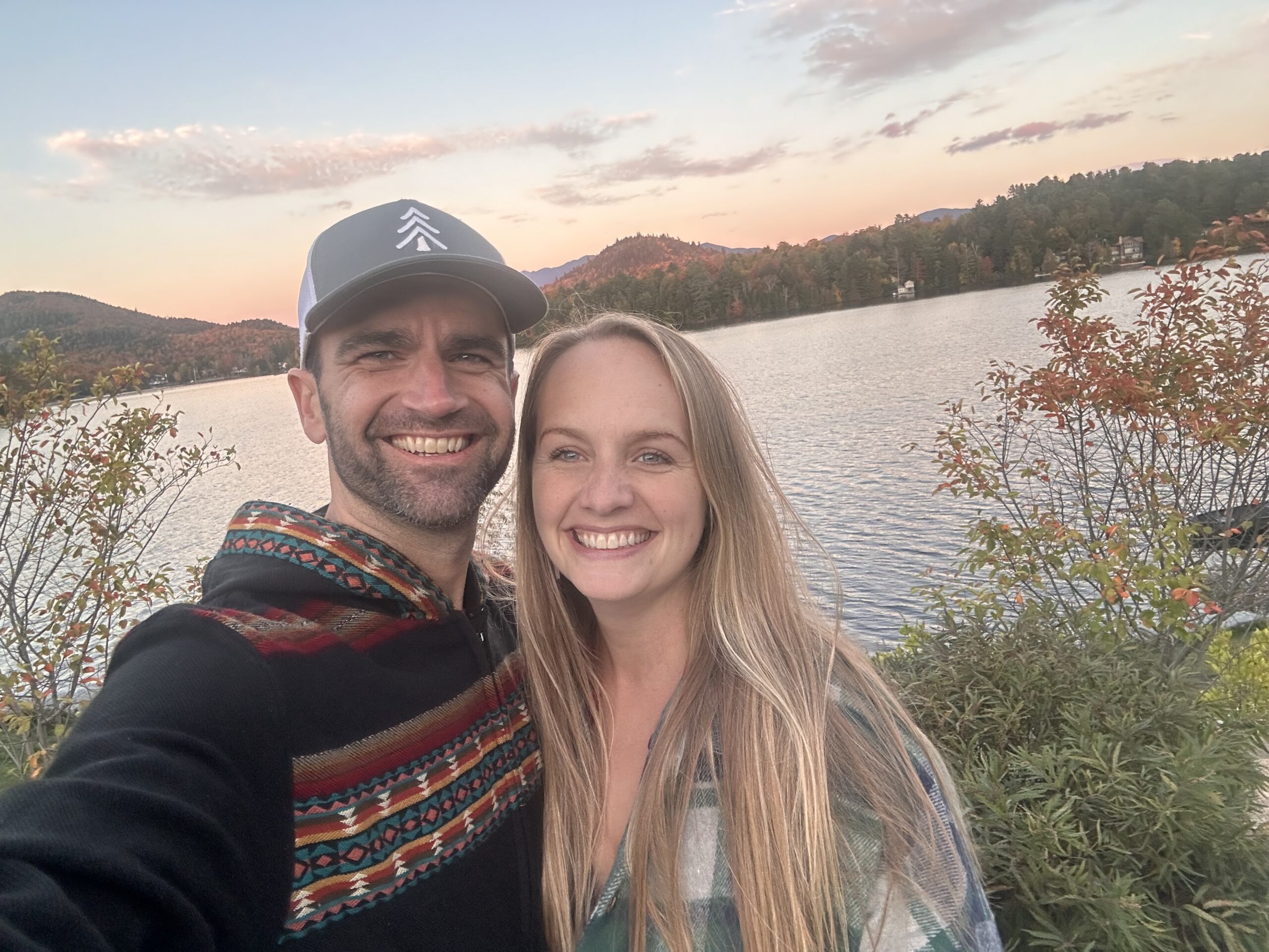 Couple smiles for a selfie by a lake at sunset with autumn foliage on the hills.