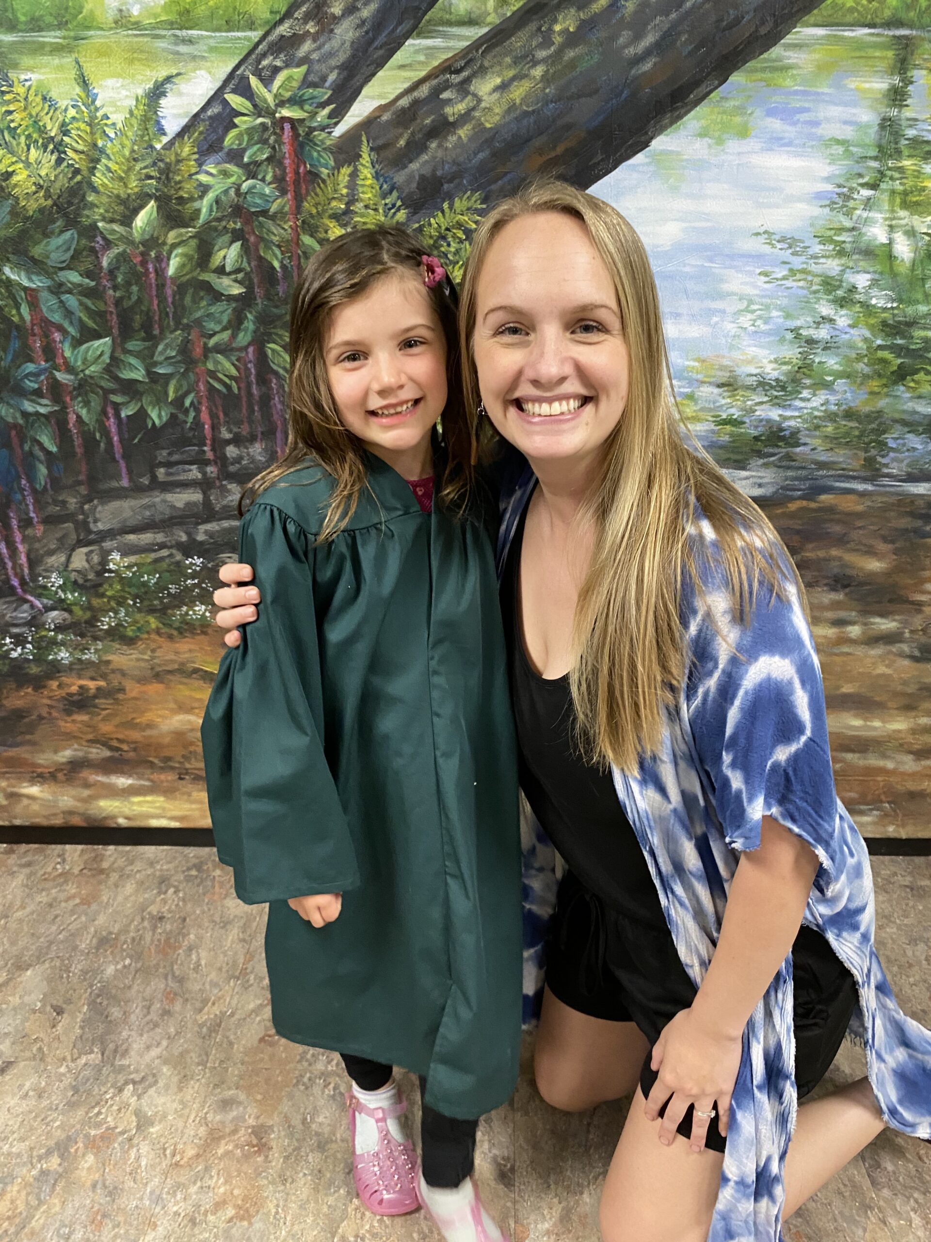A woman in a tie-dye cover-up hugs a child in a green graduation gown on a stone floor.